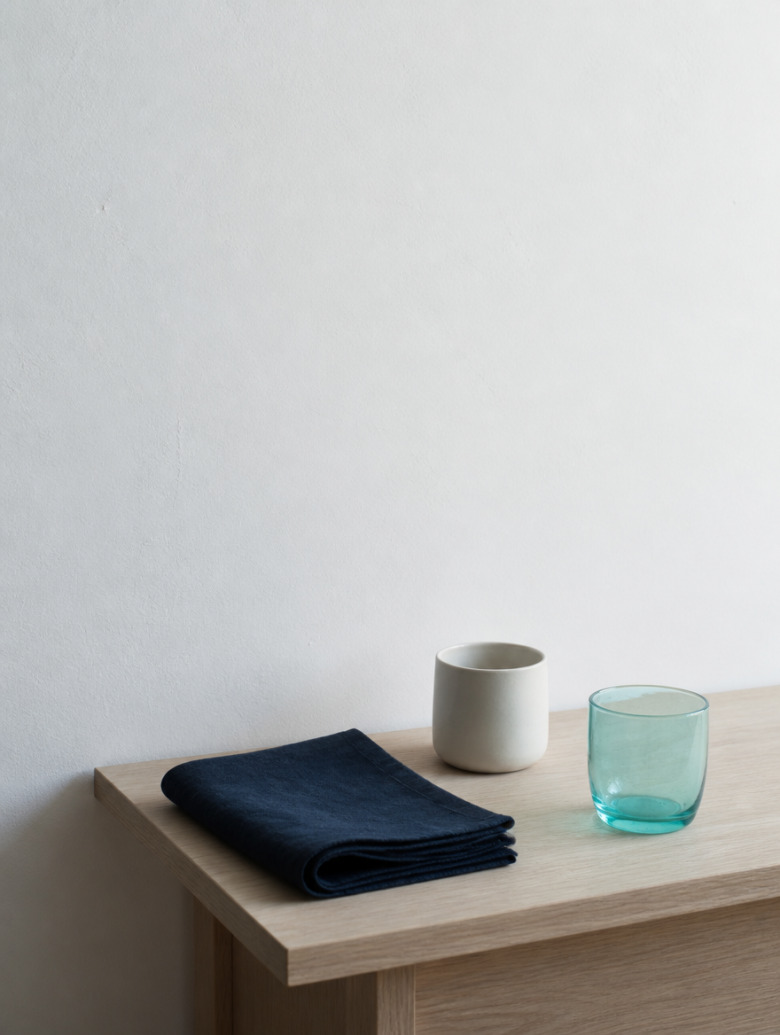 A domestic corner in morning light: a light wooden table, a folded navy cloth, a ceramic cup and an aqua glass tumbler.